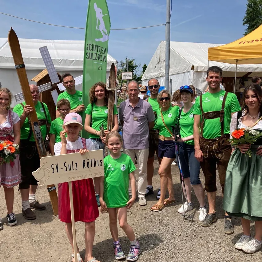 Eine Gruppe von Menschen in grünen Shirts und Lederhosen posiert für ein Foto mit einem Holzschild, auf dem 'SV-Sulzer Rothis' steht. Sie stehen draußen mit Zelten, einer Windmühle und Bäumen im Hintergrund.