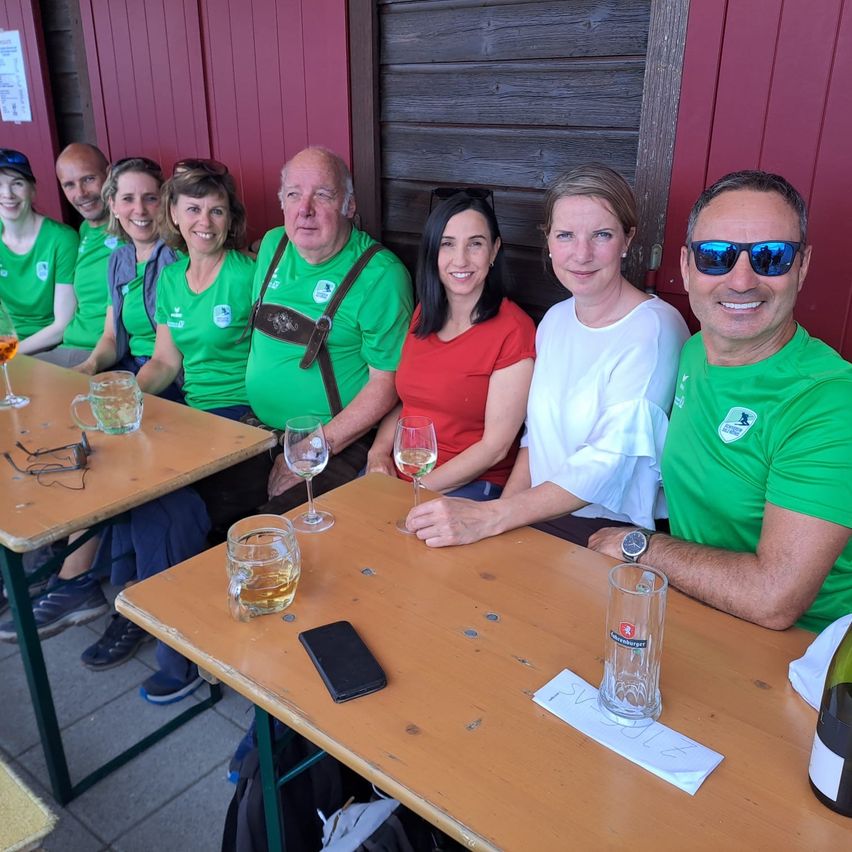 Eine Gruppe von Menschen in Grün sitzt an einem Tisch und lächelt für ein Foto. Zwei Weingläser, eine Flasche und ein Handy stehen auf dem Tisch. Dahinter ist eine rote Wand.