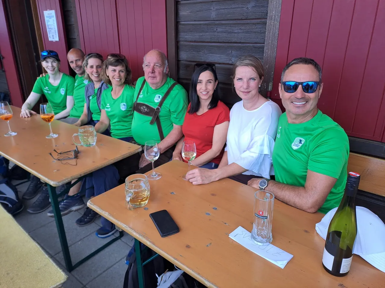 Eine Gruppe von Menschen in Grün sitzt an einem Tisch und lächelt für ein Foto. Zwei Weingläser, eine Flasche und ein Handy stehen auf dem Tisch. Dahinter ist eine rote Wand.