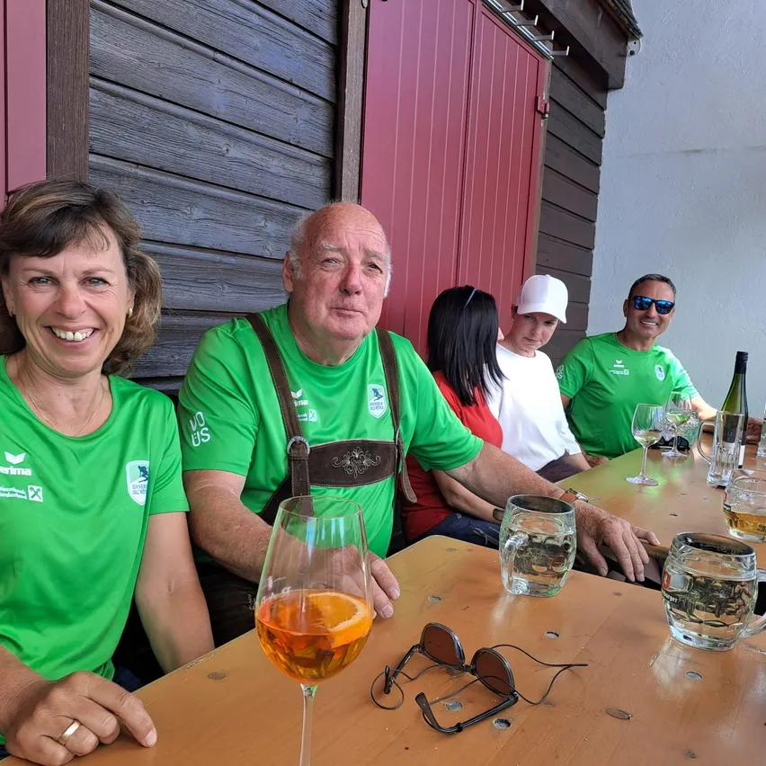 Fünf Personen sitzen lächelnd an einem Tisch und tragen grüne T-Shirts. Ein Mann trägt Hosenträger, und auf dem Tisch befinden sich Weingläser, eine Tasse und eine Flasche. Hinter ihnen steht eine rote Tür und eine Holzwand.