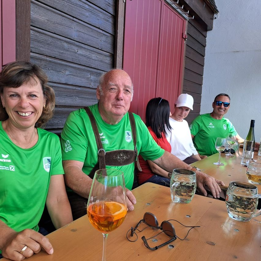 Fünf Personen sitzen lächelnd an einem Tisch und tragen grüne T-Shirts. Ein Mann trägt Hosenträger, und auf dem Tisch befinden sich Weingläser, eine Tasse und eine Flasche. Hinter ihnen steht eine rote Tür und eine Holzwand.