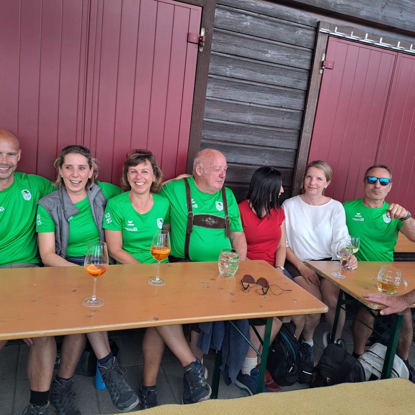 Eine Gruppe von Menschen in grünen Shirts sitzt auf Bänken mit Tischen davor, wahrscheinlich in einem Restaurant. Sie halten Gläser Bier in der Hand und posieren für ein Foto.