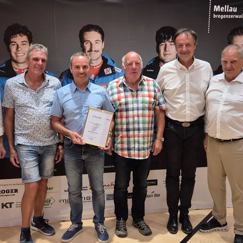 Fünf Männer stehen zusammen, einer hält eine Urkunde. Sie stehen vor einem großen Banner mit Bildern von Männern. Sie lächeln und posieren für ein Foto.