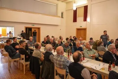 Eine große Gruppe von Menschen, die um Tische in einem Raum sitzen, einige tragen Hüte und Brillen, mit Flaschen und Papieren auf den Tischen, und zwei Personen stehen davor.
