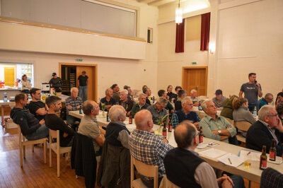 Eine große Gruppe von Menschen, die um Tische in einem Raum sitzen, einige tragen Hüte und Brillen, mit Flaschen und Papieren auf den Tischen, und zwei Personen stehen davor.
