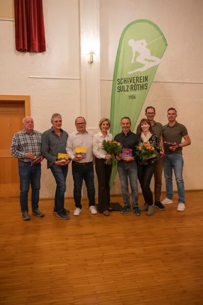 Mehrere Menschen stehen und posieren für ein Foto in einem Raum. Sie halten Blumen und Auszeichnungen. Eine grüne Fahne mit einem Skifahrer und dem Text 'SCHIVEREI SULZ-ROTHIS 1924' hängt an der Wand.