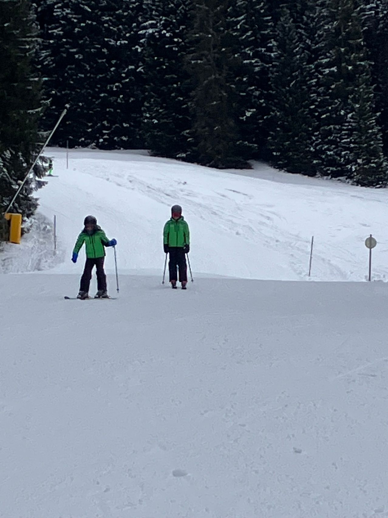 Zwei Personen steigen einen verschneiten Hang hinunter. Beide tragen grüne Jacken und halten Skistöcke. Die Person vorne trägt einen Helm.