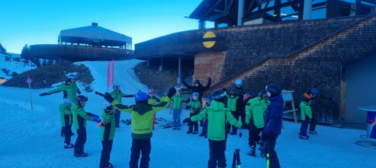 Eine Gruppe von Kindern in grüner Skiausrüstung steht auf einem verschneiten Hügel, einige heben die Hände. Dahinter ein Gebäude mit einem gelben Logo und einer roten Fahne. In der Ferne ein blauer Himmel.