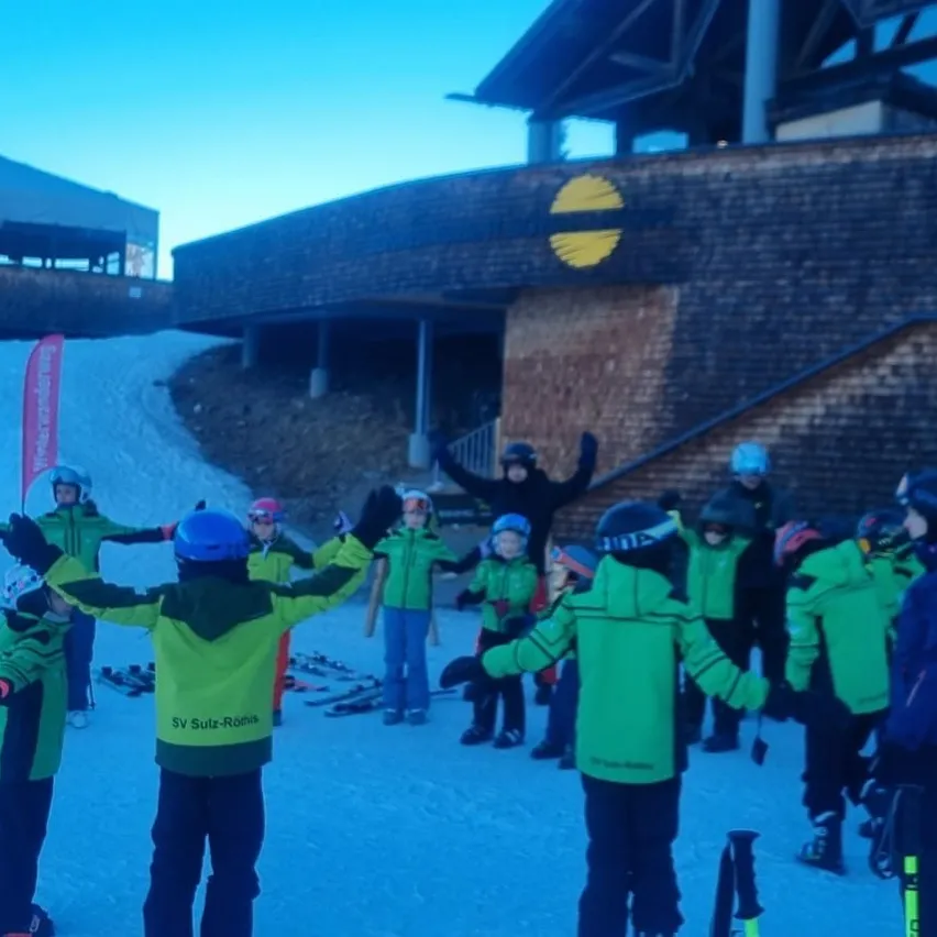 Eine Gruppe von Kindern in grüner Skiausrüstung steht auf einem verschneiten Hügel, einige heben die Hände. Dahinter ein Gebäude mit einem gelben Logo und einer roten Fahne. In der Ferne ein blauer Himmel.