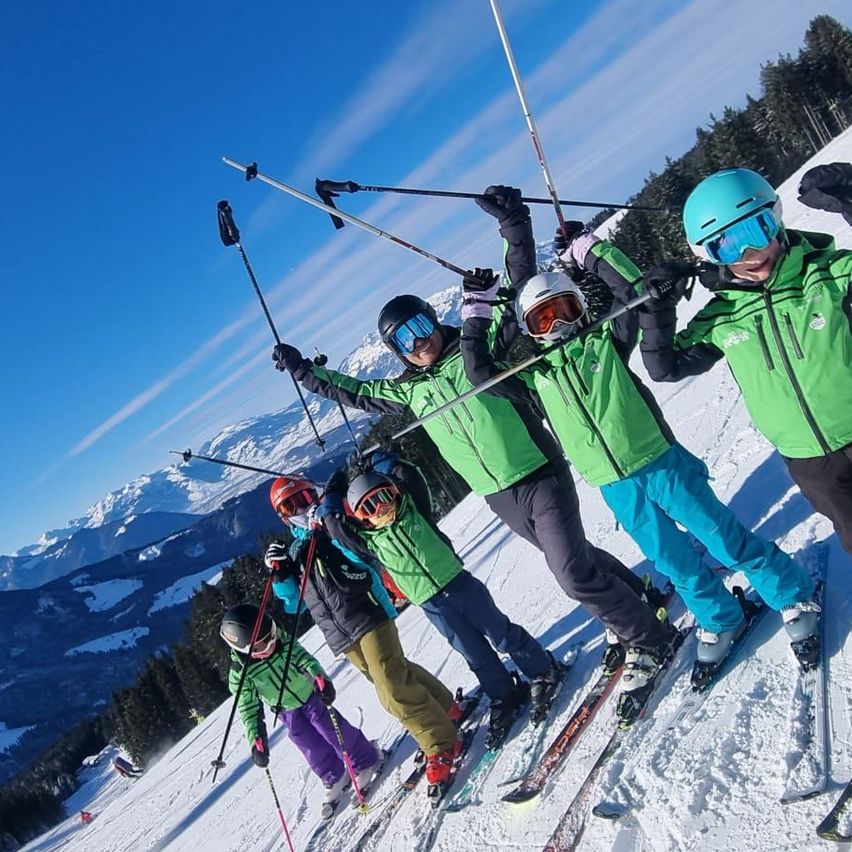 Eine Gruppe von Menschen skifahrert auf einem Berg und trägt grüne Jacken, Helme und Skischuhe. Sie halten Skistöcke. Der Berg hat Bäume und Schnee.