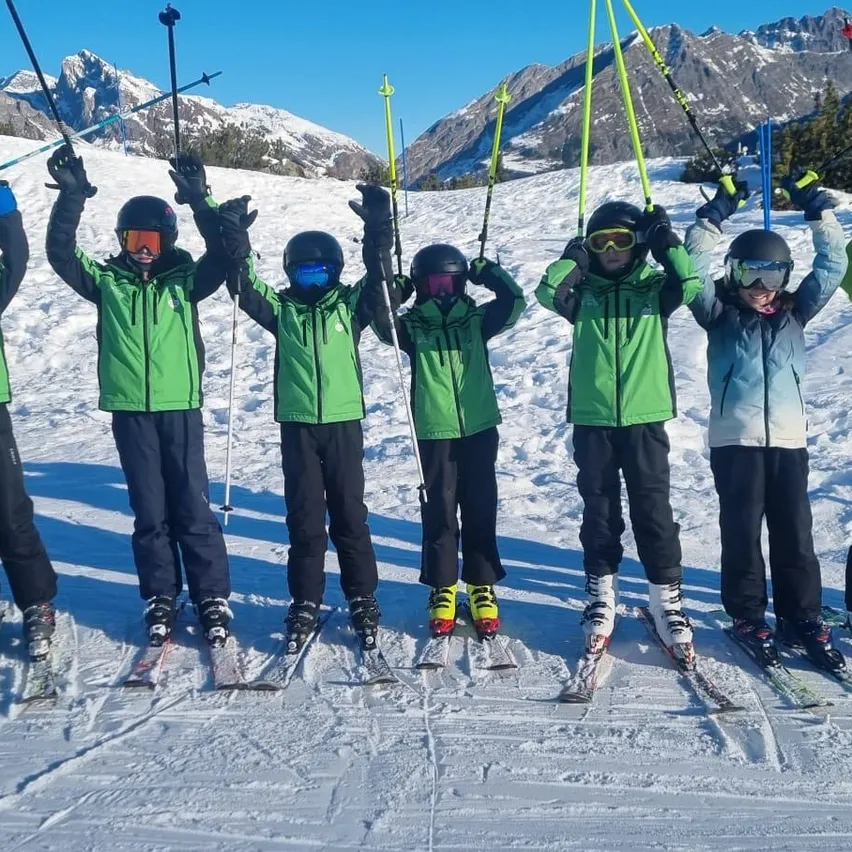 Sechs Skifahrer in grünen und schwarzen Skianzügen, Helmen und Schutzbrillen posieren für ein Foto auf einem verschneiten Berg.