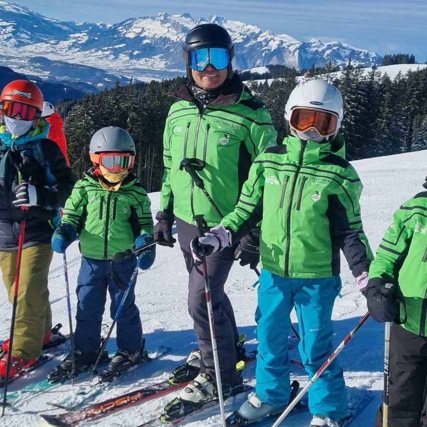 Eine Gruppe von Menschen, darunter Erwachsene und Kinder, steht auf einem verschneiten Berg und trägt alle grüne Skijacken und Helme. Sie halten Skistöcke in der Hand und lächeln in die Kamera.