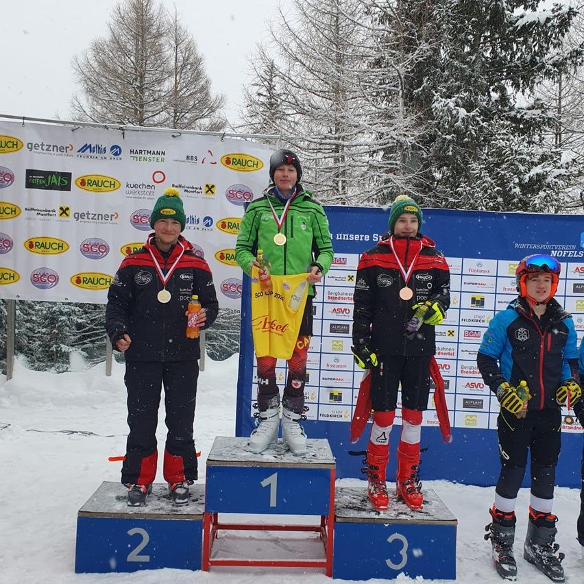 Vier Kinder stehen auf einem Podest im Schnee. Sie tragen Skischuhe und Jacken. Sie haben Medaillen um den Hals. Das Podest hat die Nummern 1, 2 und 3 darauf. Hinter ihnen ist ein Banner mit Logos.