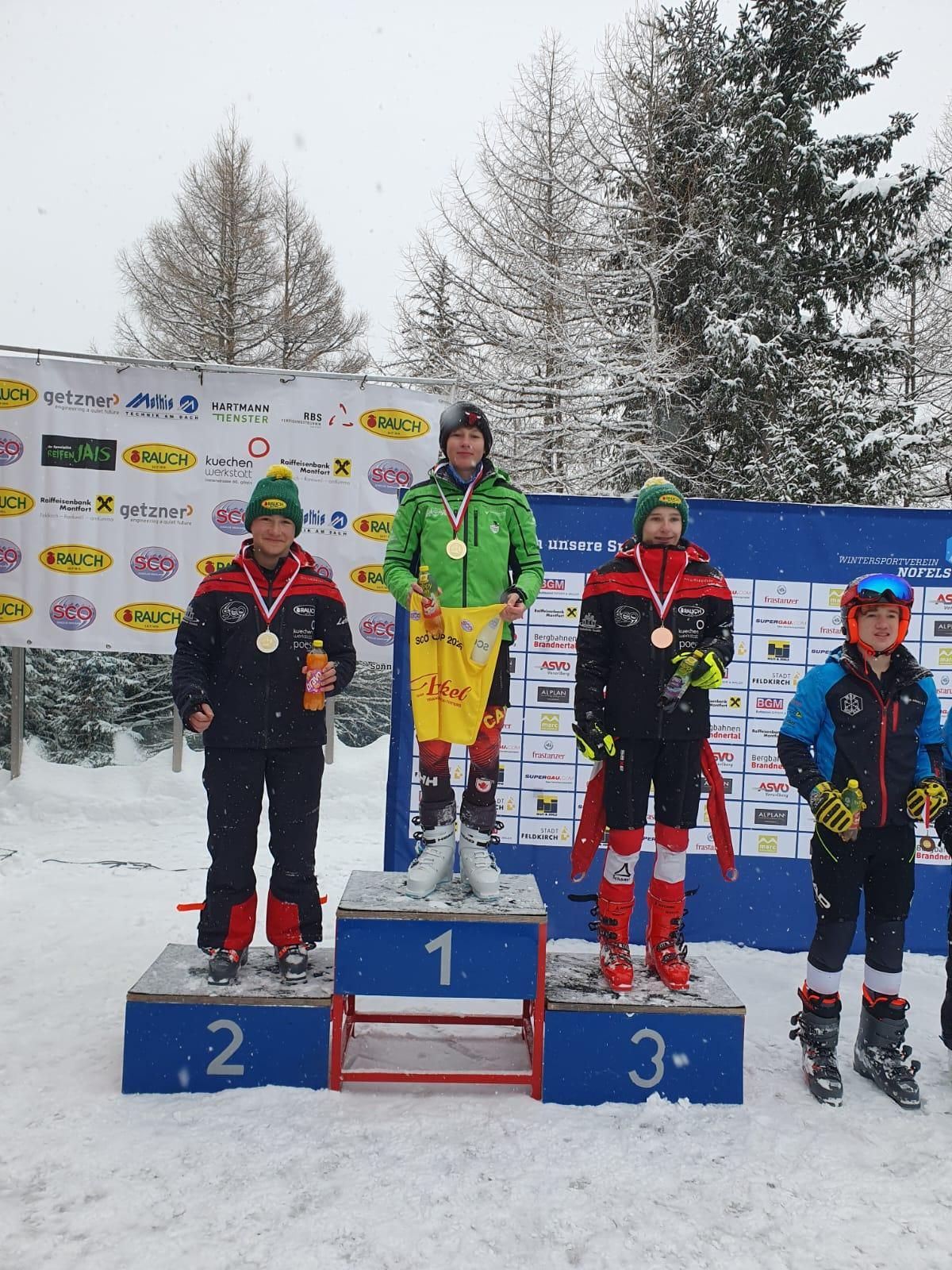 Vier Kinder stehen auf einem Podest im Schnee. Sie tragen Skischuhe und Jacken. Sie haben Medaillen um den Hals. Das Podest hat die Nummern 1, 2 und 3 darauf. Hinter ihnen ist ein Banner mit Logos.