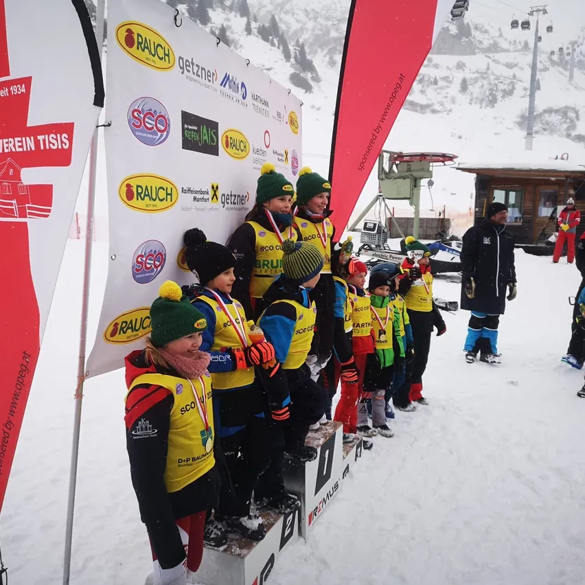 Eine Gruppe von Kindern in gelber und blauer Skiausrüstung steht auf einem Podest im Schnee, trägt Medaillen und Mützen. Hinter ihnen ist ein Banner mit Logos und Text zu sehen. Ein Mann in einem schwarzen Mantel steht im Hintergrund.