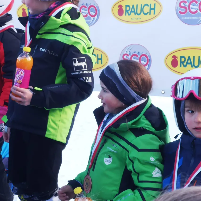 Mehrere Kinder in grünen und schwarzen Skijacken mit Medaillen stehen im Schnee, eines hält eine Flasche Yazoo. Hinter ihnen ist eine Wand mit Logos wie RAU und SCO.