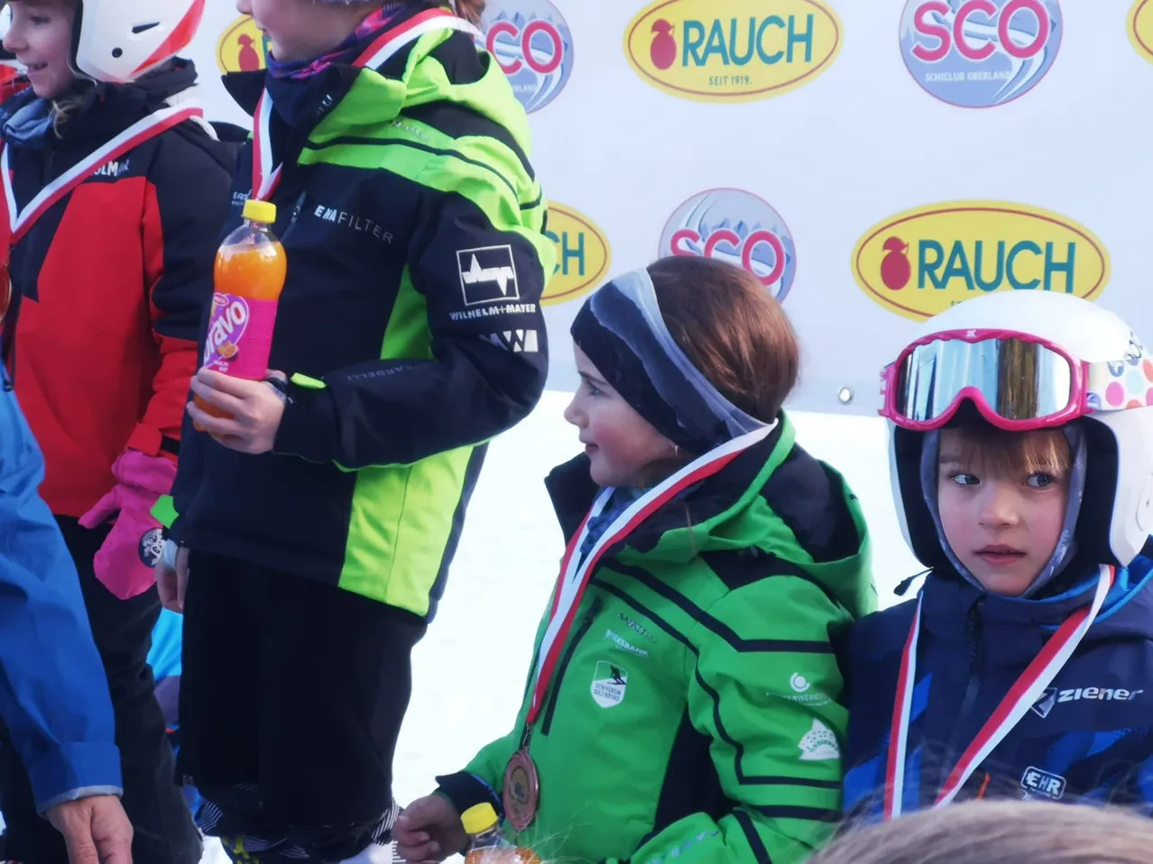 Mehrere Kinder in grünen und schwarzen Skijacken mit Medaillen stehen im Schnee, eines hält eine Flasche Yazoo. Hinter ihnen ist eine Wand mit Logos wie RAU und SCO.