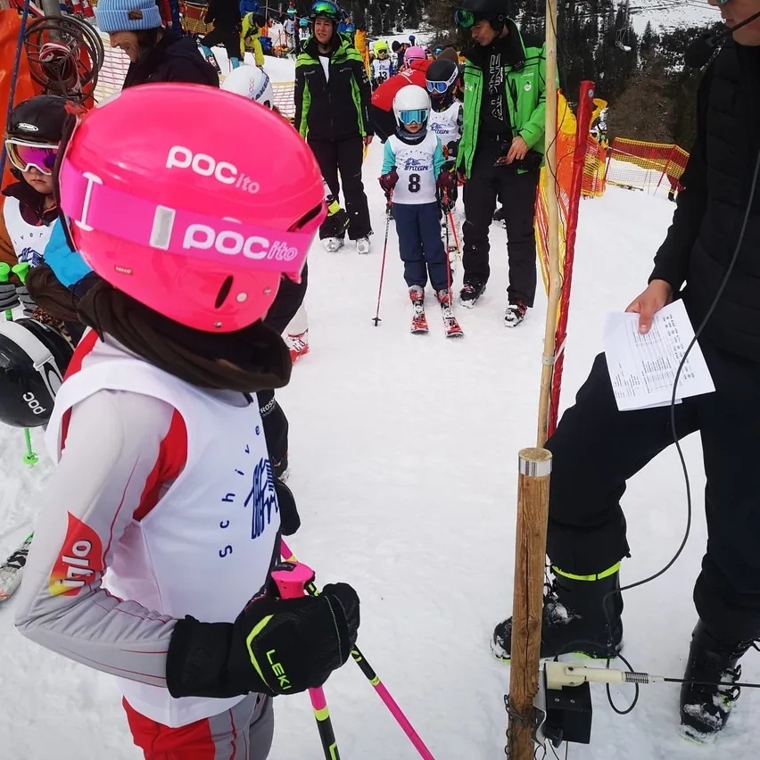 Mehrere Kinder und Erwachsene sind auf einer verschneiten Piste Skifahren. Ein Kind mit einem rosa Helm und einer Schneebrille schaut in die Kamera. Ein weiteres Kind mit einem weißen Helm und Trikot mit der Nummer 8 skis mit einem Trainer.