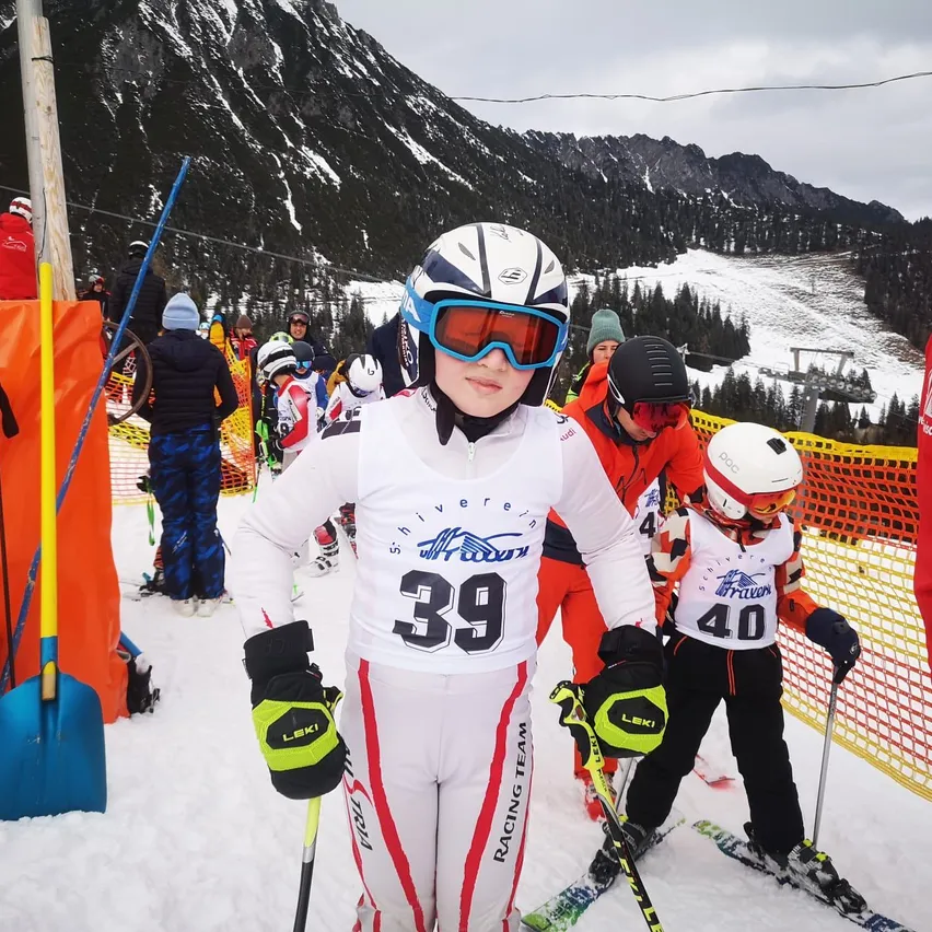 Ein junger Skifahrer in einem weißen und roten Skianzug mit der Nummer 39 steht auf einem verschneiten Hang. In der Nähe bereiten sich andere Skifahrer, einige in orangefarbenen Anzügen, auf ein Rennen vor. Im Hintergrund sind Berge und eine Skiliftanlage zu sehen.