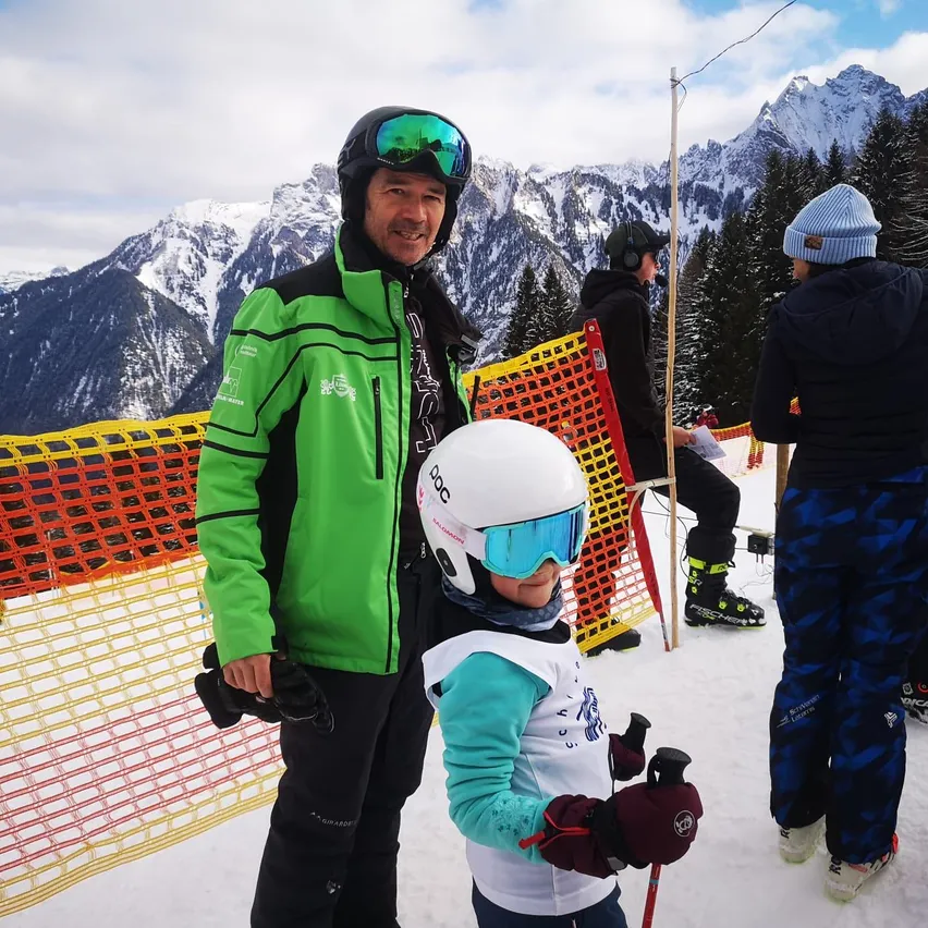 Ein Mann und ein Kind sind auf einem verschneiten Berg beim Skifahren. Im Hintergrund sind Bäume und Berge zu sehen. Das Kind trägt einen Helm und eine Schutzbrille.