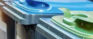 Vier Recyclingbehälter mit verschieden farbigen Deckeln sind aufgereiht. Der erste ist blau, der zweite schwarz mit rotem und weißem Design, der dritte grün und der letzte schwarz.