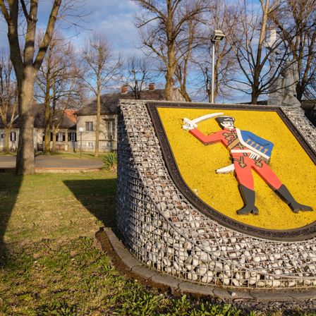 Ein gelber Schild mit einer Soldatenfigur darauf steht auf einem Rasen, mit Gebäuden und Bäumen im Hintergrund.