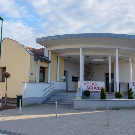 Das Volksschule Nepomuk Gebäude hat ein geschwungenes Dach und weiße Säulen, eine Treppe und einen Garten. Ein Schild steht 'VOLKS-SCHULE NEPOMUK'.