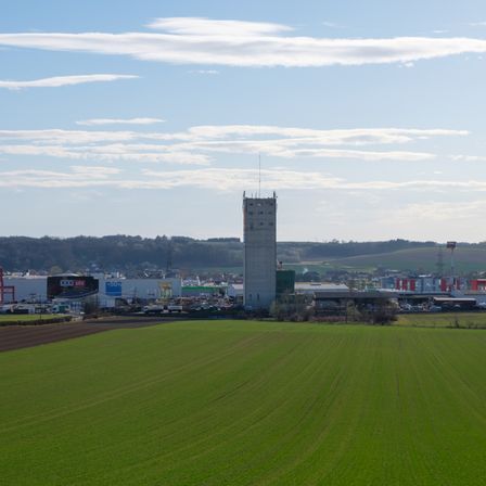 Ein breites grünes Feld mit einem hohen Turm in der Ferne und mehreren Gebäuden. Bäume und Berge sind im Hintergrund sichtbar.