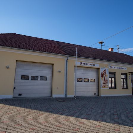 Ein gelbes Gebäude mit braunem Dach hat zwei weiße Garagen mit kleinen Fenstern, einen Fahnenmast und ein Schild mit der Aufschrift 'Feuerwehr'. An der Wand hängt ein Poster einer Frau.