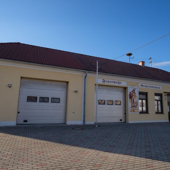Ein gelbes Gebäude mit braunem Dach hat zwei weiße Garagen mit kleinen Fenstern, einen Fahnenmast und ein Schild mit der Aufschrift 'Feuerwehr'. An der Wand hängt ein Poster einer Frau.