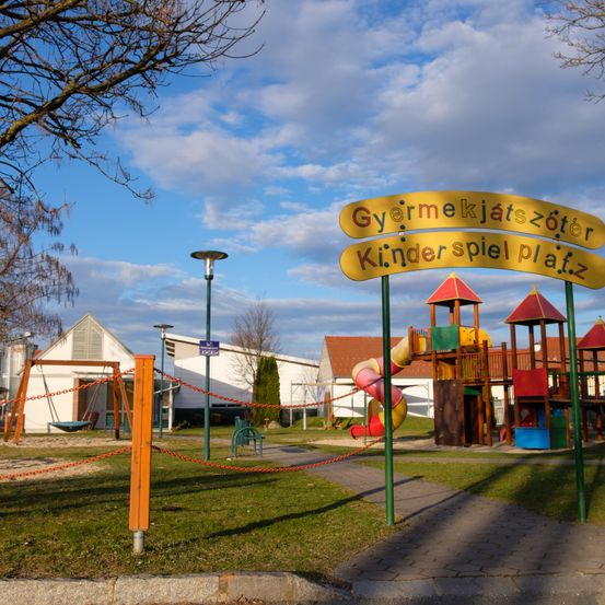 Ein Spielplatz mit einer Rutsche und Schaukeln unter einem blauen Himmel mit Wolken. Das Schild sagt 'Gyermekjátszóter Kinder spiel platz.'