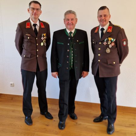 Drei Männer stehen zusammen in einem Raum. Die beiden an den Seiten tragen Militäruniformen mit Medaillen. Der in der Mitte trägt einen schwarzen Anzug mit einer grünen Weste.