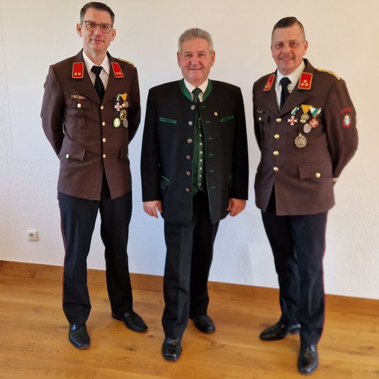 Drei Männer stehen zusammen in einem Raum. Die beiden an den Seiten tragen Militäruniformen mit Medaillen. Der in der Mitte trägt einen schwarzen Anzug mit einer grünen Weste.