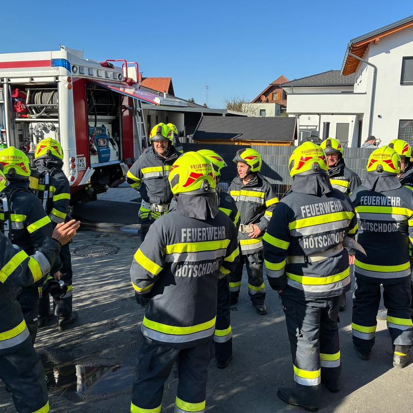 Eine Gruppe Feuerwehrleute in gelben und schwarzen Uniformen steht vor einem Feuerwehrauto, mit Häusern dahinter.