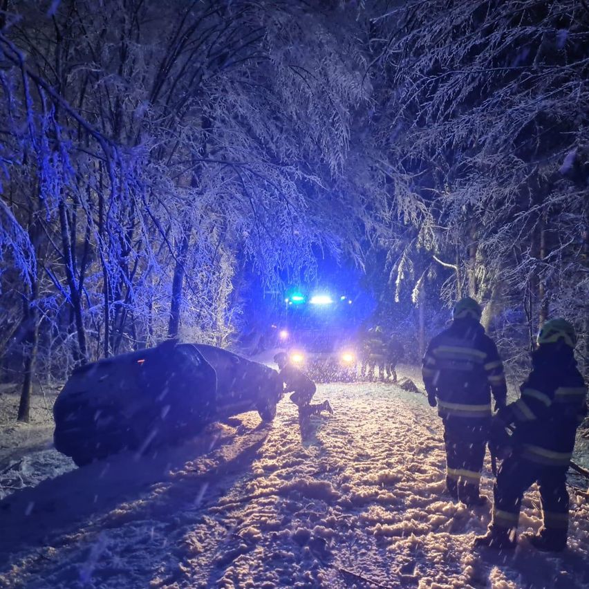 Feuerwehrleute in Uniform helfen einer Person in der Nähe eines Autos im Schnee. Das Gebiet ist dunkel, mit Bäumen bedeckt von Eis und einem Polizeiauto mit blinkenden blauen Lichtern in der Nähe.