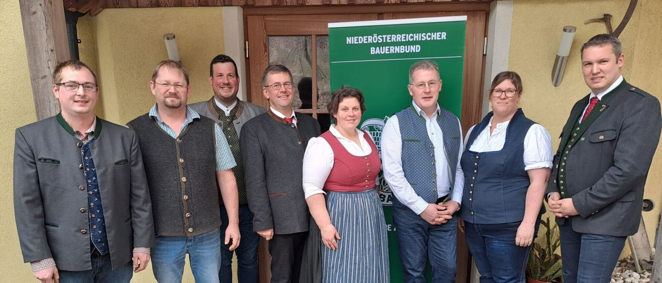 Eine Gruppe von sechs Personen steht vor einem grünen Banner mit weißen Text. Der Mann links trägt ein blaues Hemd und eine Brille. Die zentrale Figur ist eine Frau in einem roten und blauen Kleid. Die anderen sind in formeller Kleidung gekleidet.