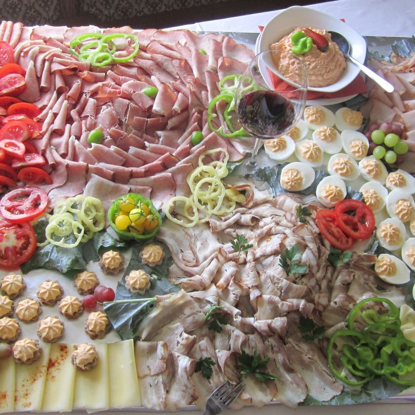Eine Platte mit verschiedenen Fleischsorten, darunter Schinken, Speck und Würstchen, begleitet von geschnittenen Tomaten, Käse, Oliven, Trauben, pochierten Eiern und einer Schüssel Dip. Ein Weinglas mit Rotwein ist ebenfalls vorhanden.