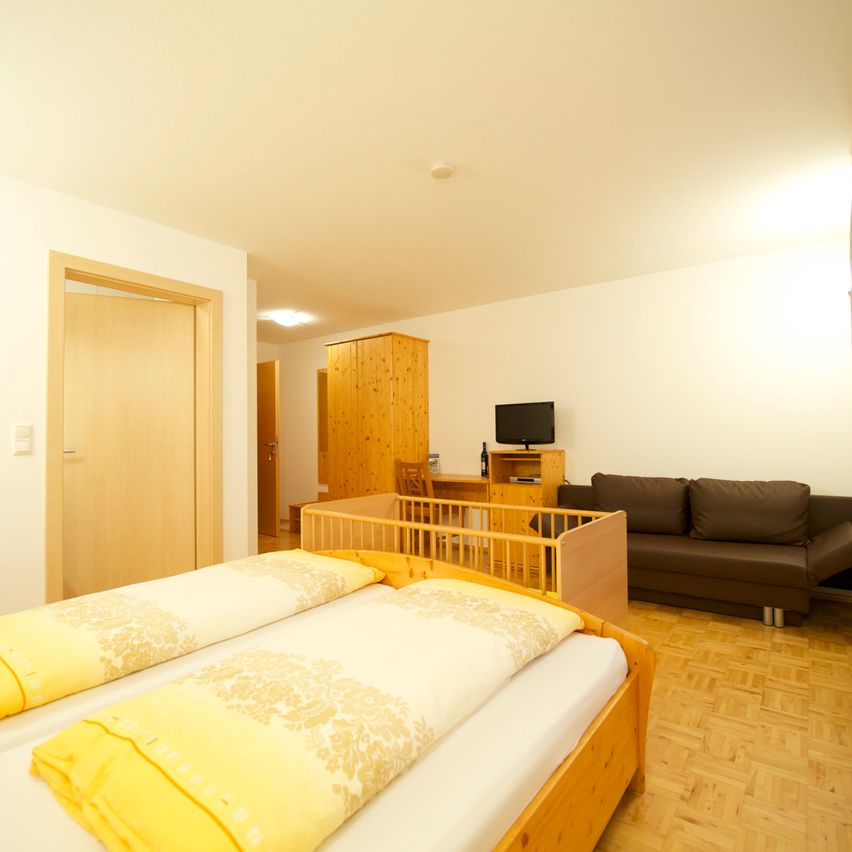 Ein gemütliches Schlafzimmer mit zwei Betten, einem hölzernen Kinderbett, einer braunen Couch, einem Schreibtisch mit Monitor, einem hölzernen Kleiderschrank und einem Holzboden.