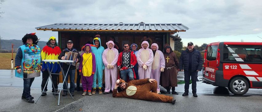 A group of people in colorful costumes pose for a photo, with one person lying on the ground in a bear costume. Behind them, there is a small building with a metal roof.