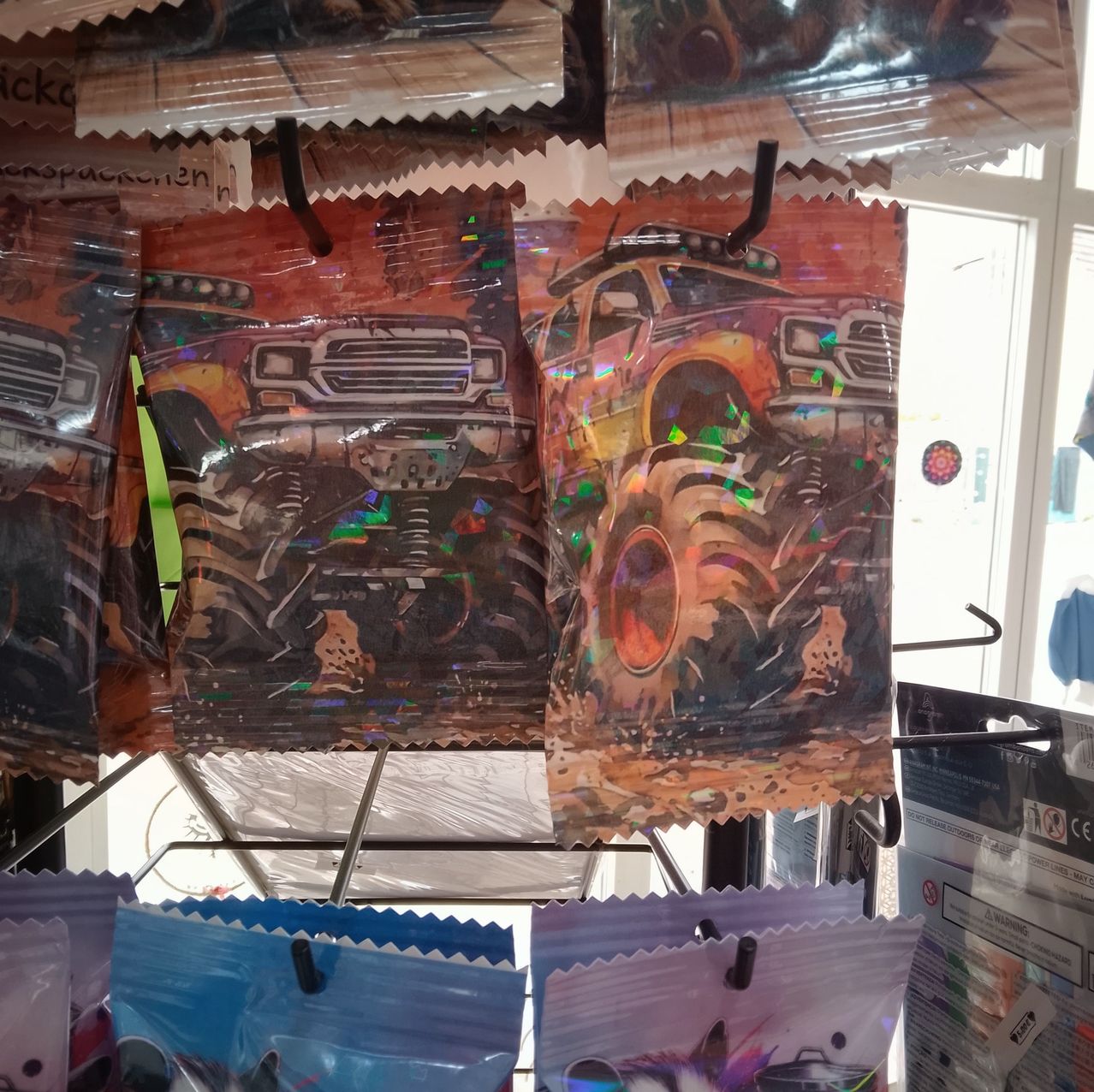 A display of colorful, holographic monster truck bags hanging on hooks in a store. The bags feature vibrant illustrations of monster trucks with large wheels and exaggerated features.