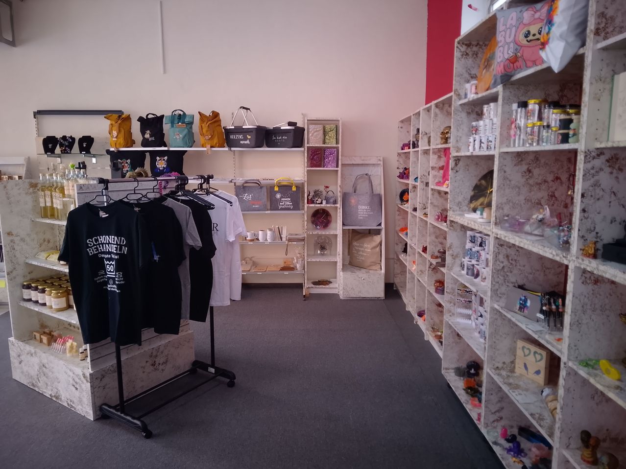 Ein Ladeninneres mit Regalen, die mit verschiedenen Artikeln gefüllt sind, darunter Taschen, Tassen und Dekorationsgegenstände. Links steht ein Ständer mit schwarzen T-Shirts. Der Boden ist mit Teppich ausgelegt, und die Wände sind in hellen Farben gestrichen.