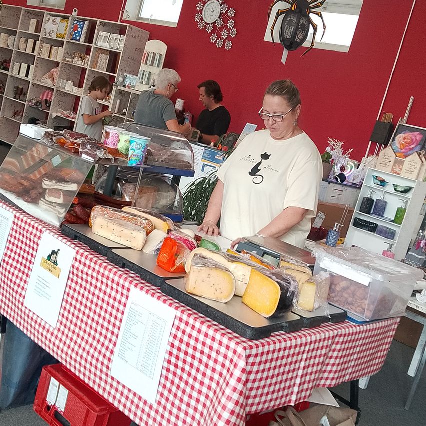 A woman stands behind a table displaying various cheeses and meats, with a red wall and shelves in the background.