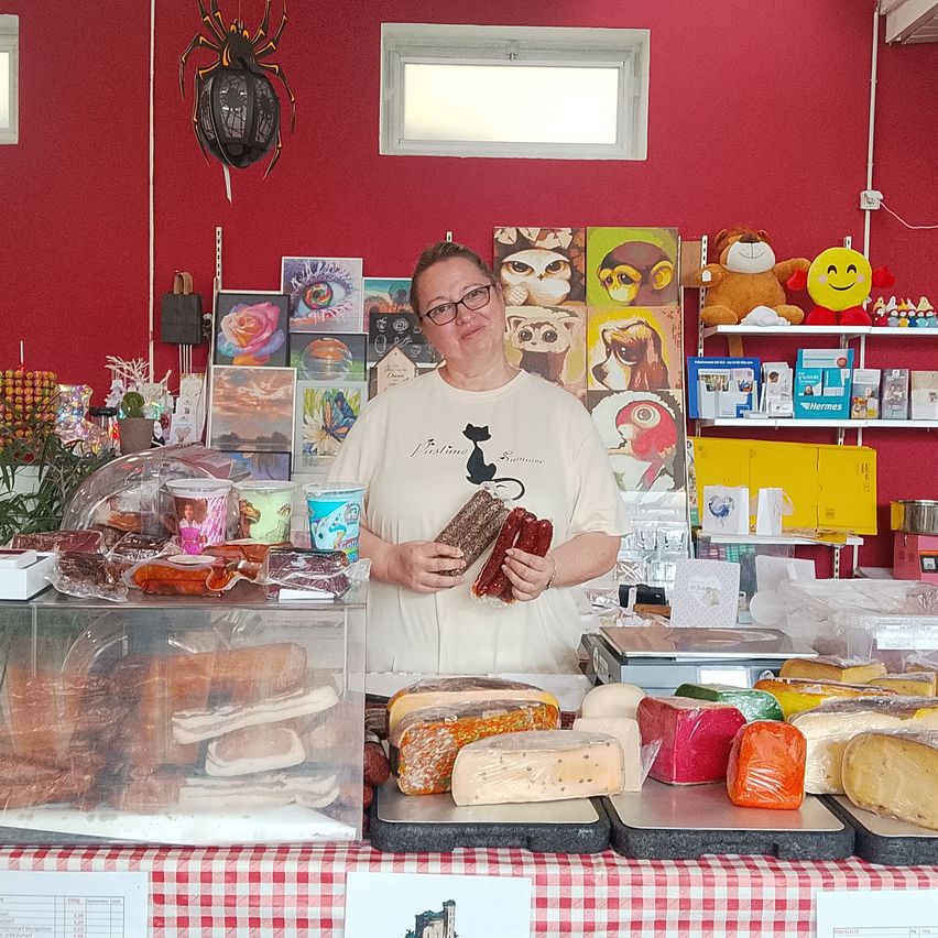 A woman stands behind a counter in a shop, holding a package of salami and smiling. The counter is filled with various meats, cheeses, and other food items. The shop has a red wall, shelves with stuffed toys, and a window above.