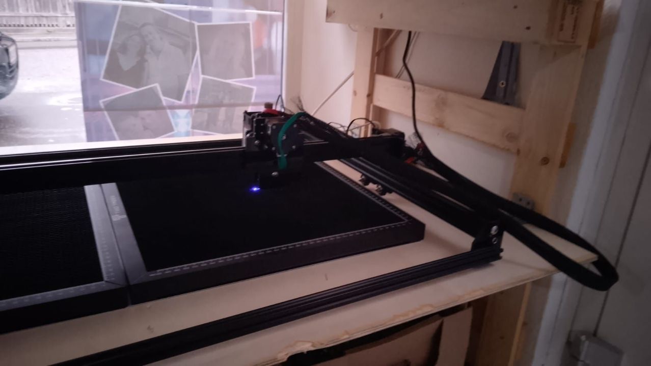 A black laser cutting machine is on a white table, with a blue light illuminating the work surface.