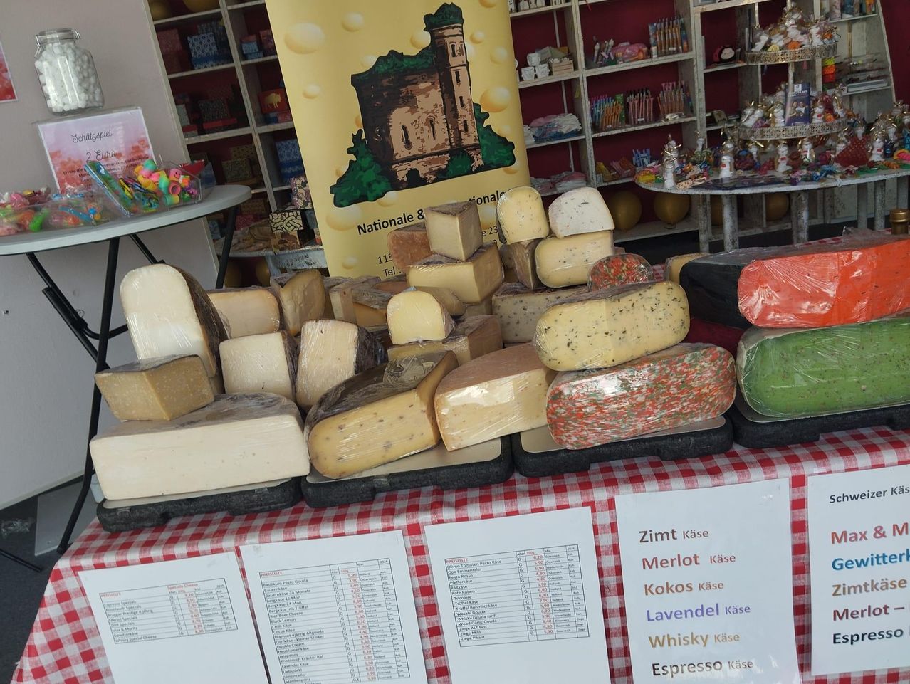 A table with a red and white tablecloth displays various cheeses with labels. Behind the table, shelves hold different items, and a yellow banner with text and an image is visible.