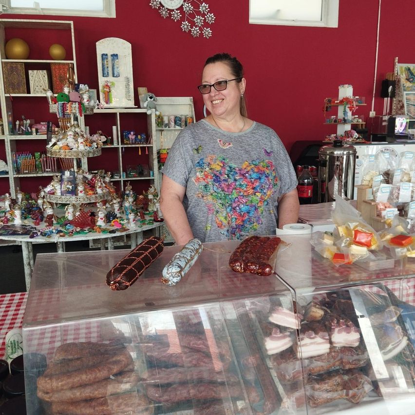 A woman stands behind a counter in a shop with various meats and desserts displayed. The counter is covered with a red-and-white tablecloth.