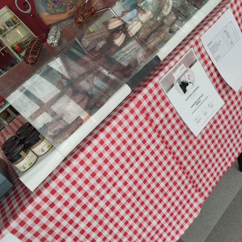 A red and white gingham table displays an assortment of sausages and cheese with a sign in front.