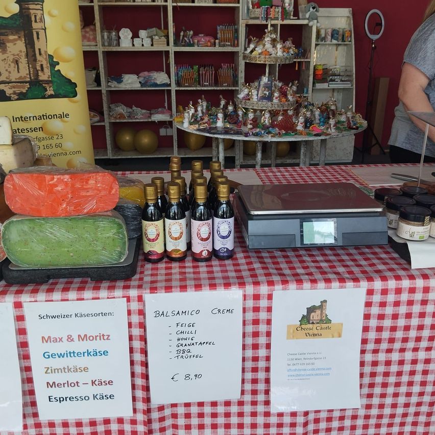 A food stall with a red and white checkered tablecloth displays various cheeses, bottles, and a scale. The cheese varieties include Max & Moritz, Gewitterkäse, Zimtkäse, Merlot-Käse, and Espresso Käse. The stall also has a price tag for €8.90. Behind it is a shelf with assorted items and a sign that reads Internationale Käsessen.