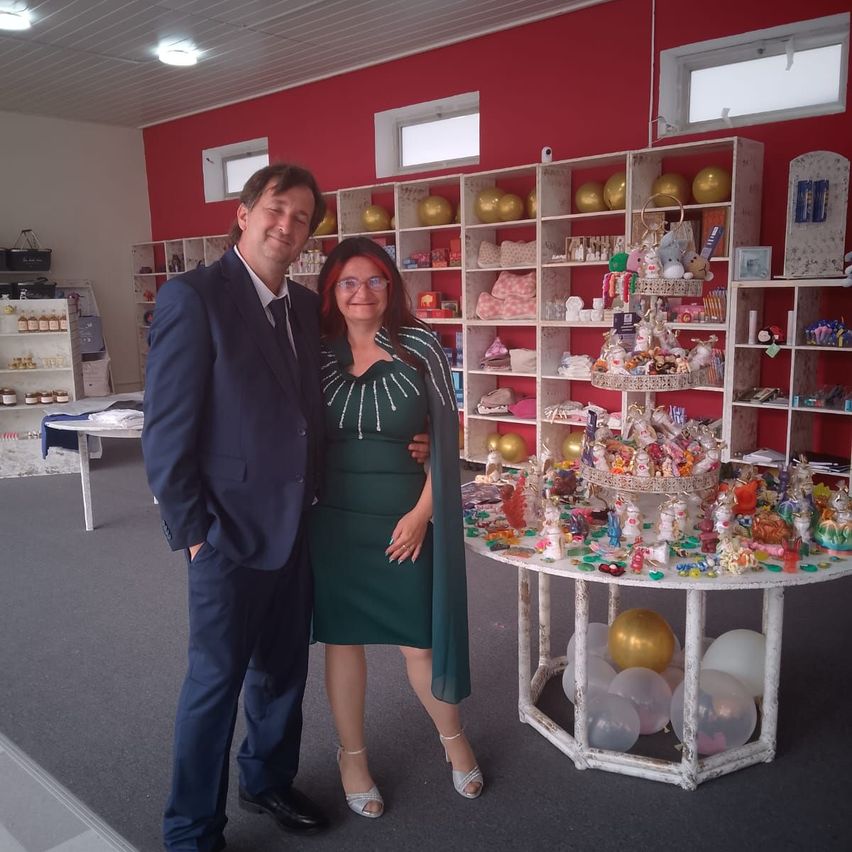 Ein Mann und eine Frau stehen neben einem Tisch mit verschiedenen Spielzeugen in einem Raum mit weißen Regalen, roten Wänden und Deckenbeleuchtung.