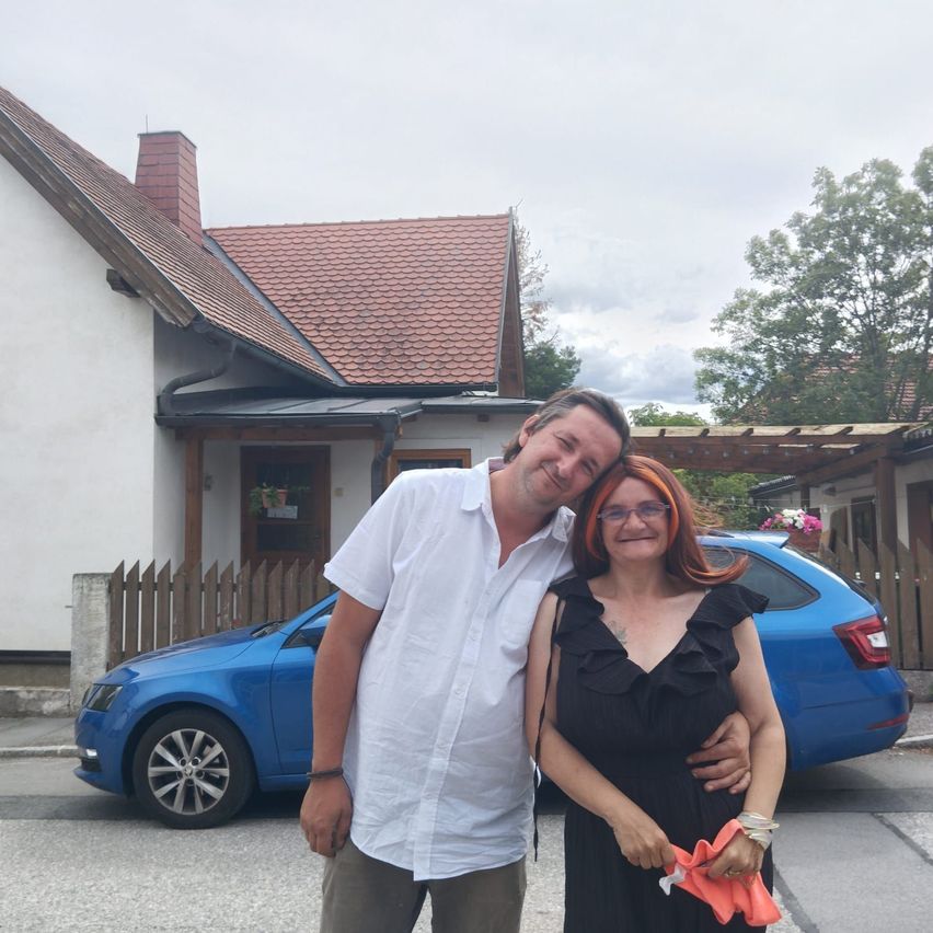 Ein Mann und eine Frau stehen vor einem Haus mit einem blauen Auto im Hintergrund. Der Mann trägt ein weißes Hemd und braune Hosen, während die Frau ein schwarzes Kleid trägt.